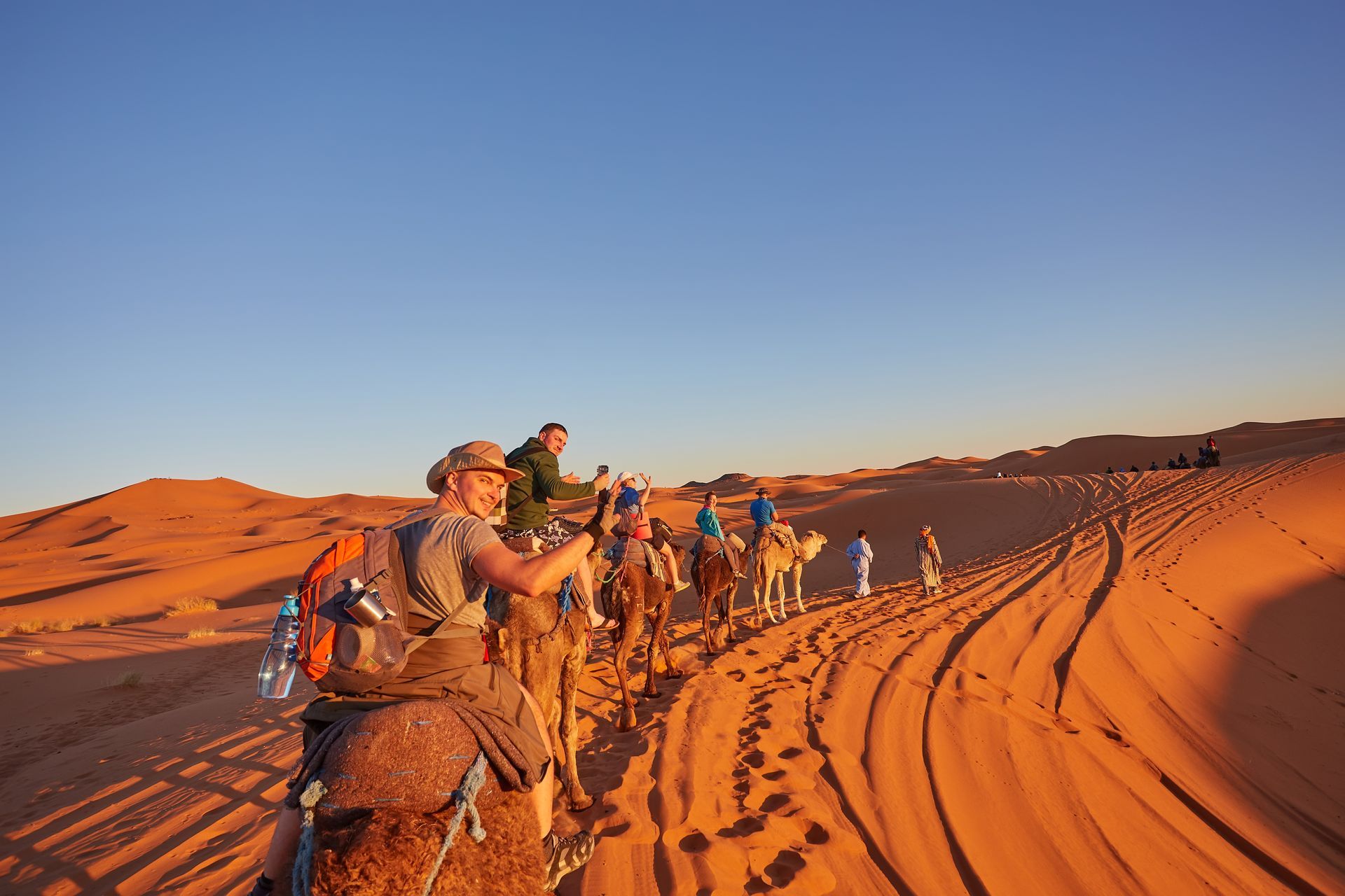 People ride camels across desert dunes under a blue sky, tracks in the sand.