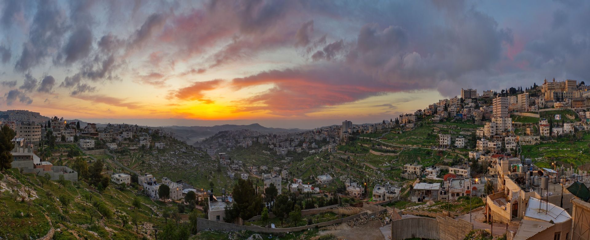 Panoramic view of a city with buildings nestled on a hillside under a colorful sunset sky.