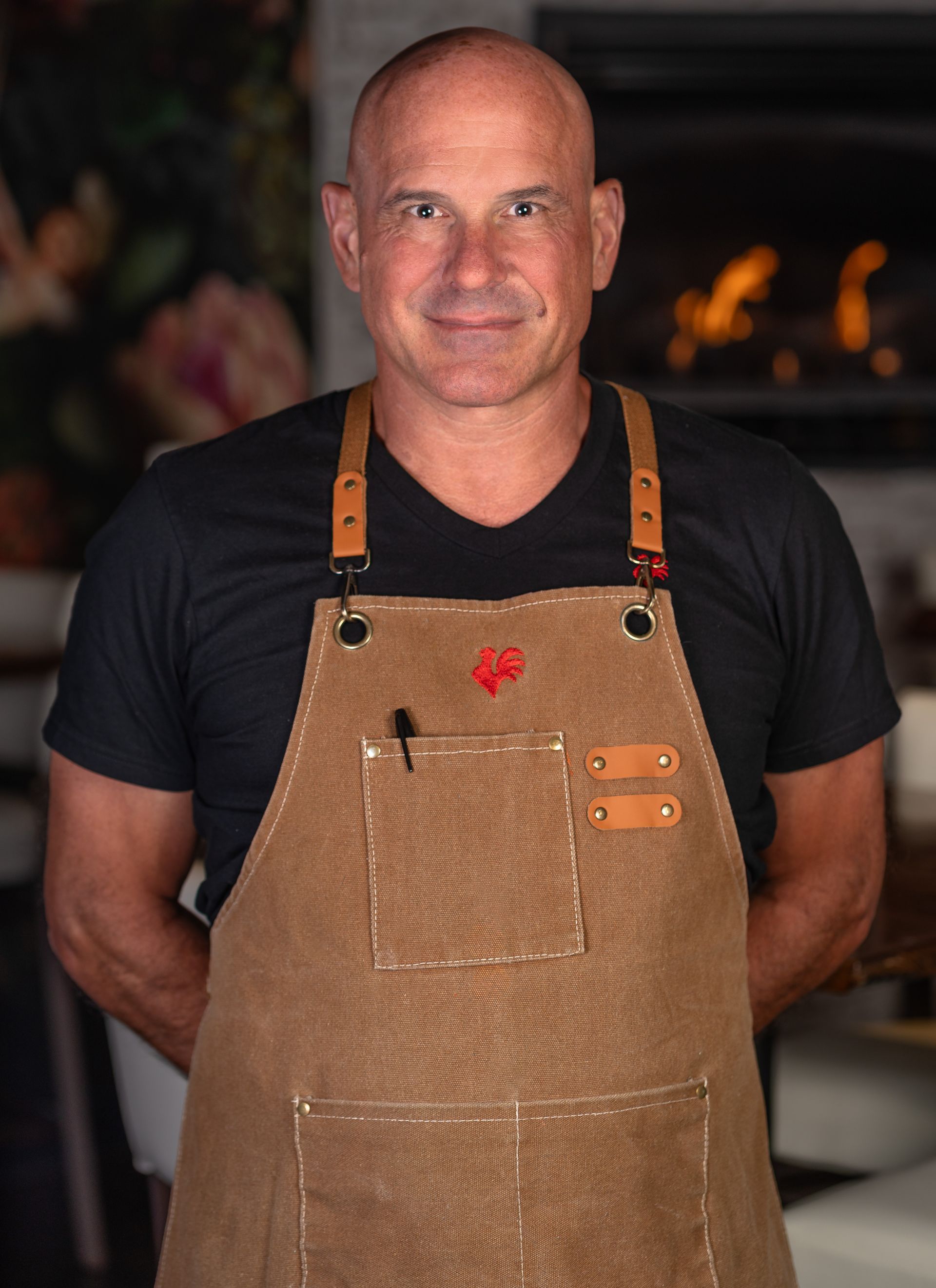 Bald man in brown apron, black shirt, smiling. Restaurant setting, fireplace visible.