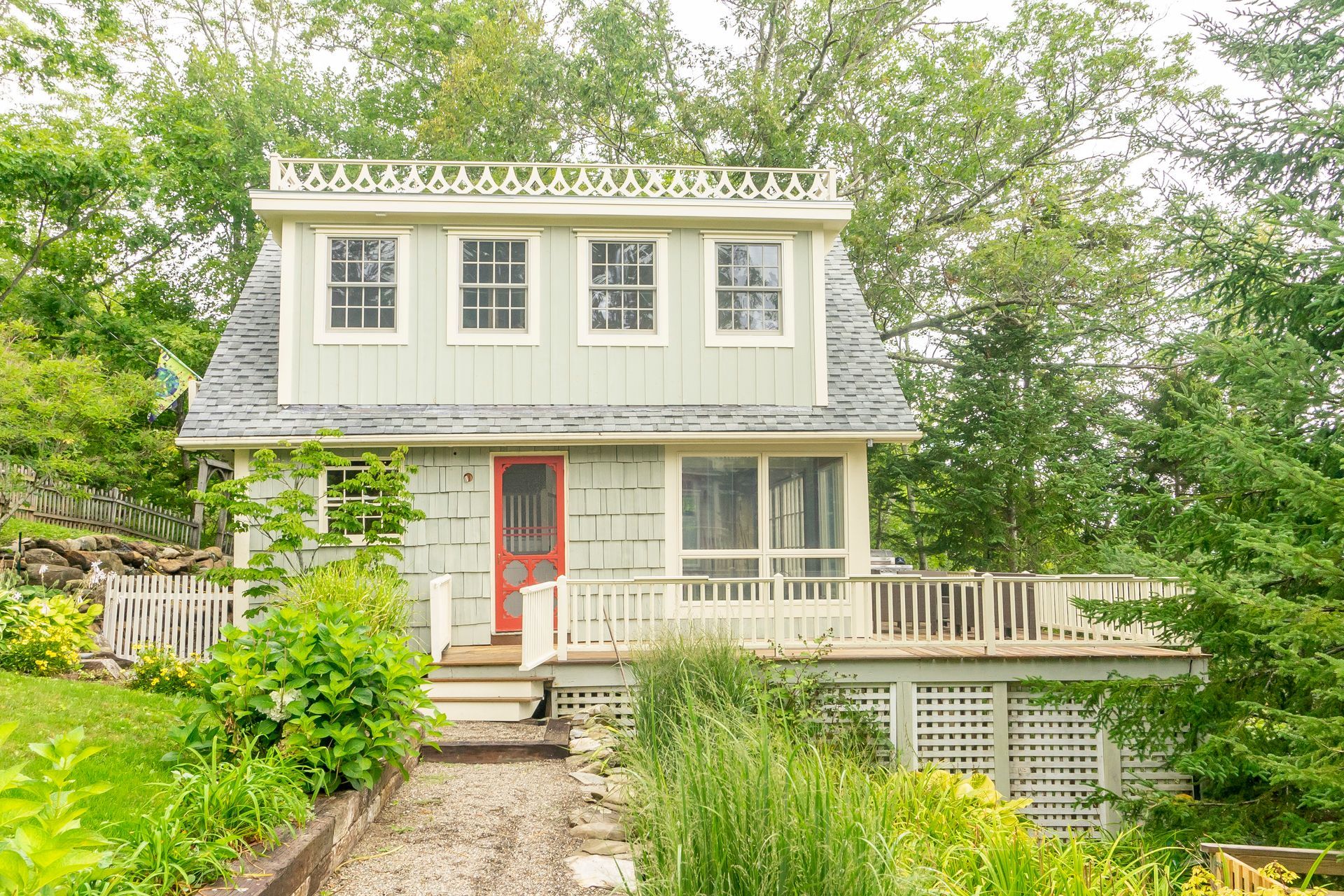 A small white house with a red door and a large deck surrounded by trees.