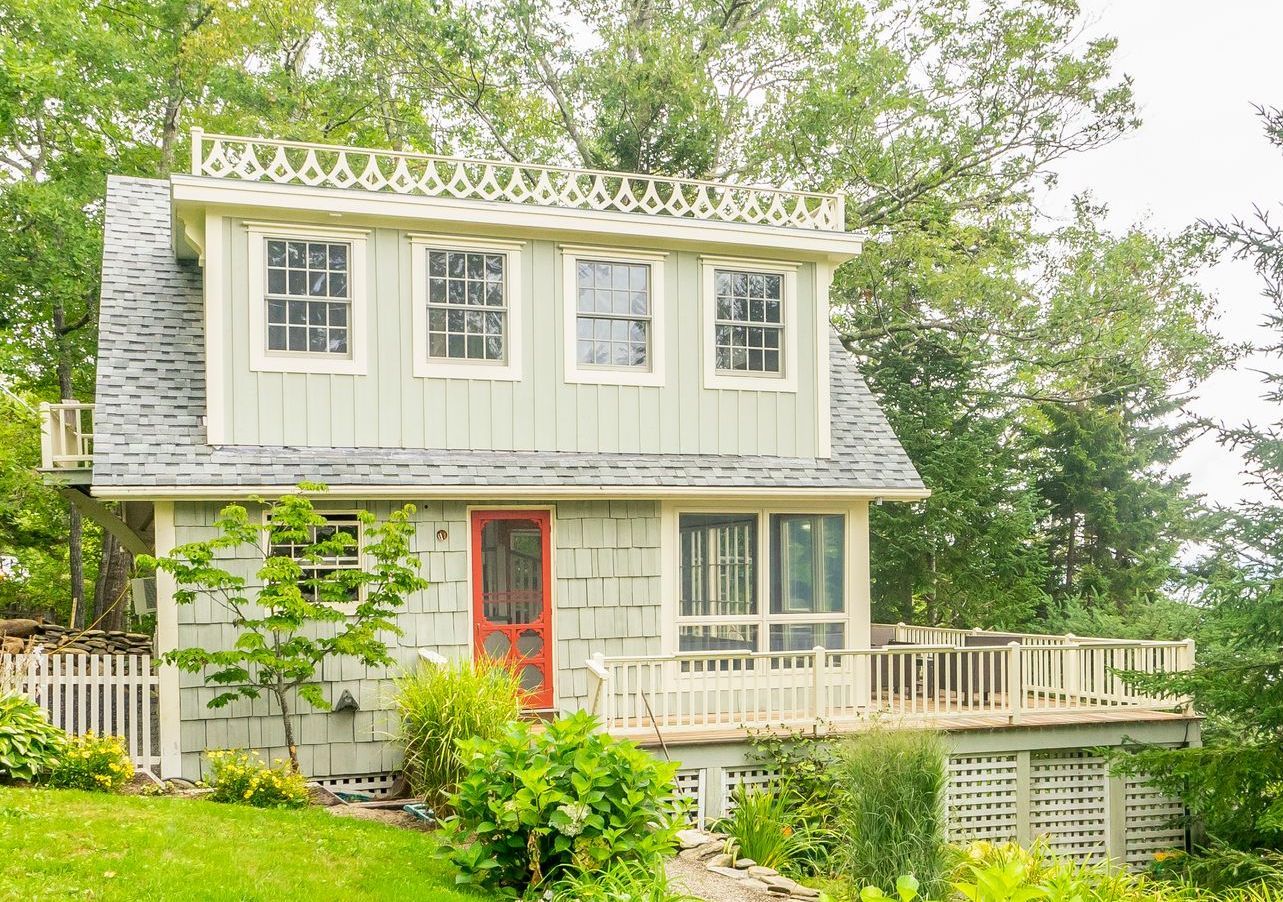 Small, two-story house with green siding, red door, and decorative trim. Wooden deck in front, surrounded by greenery.