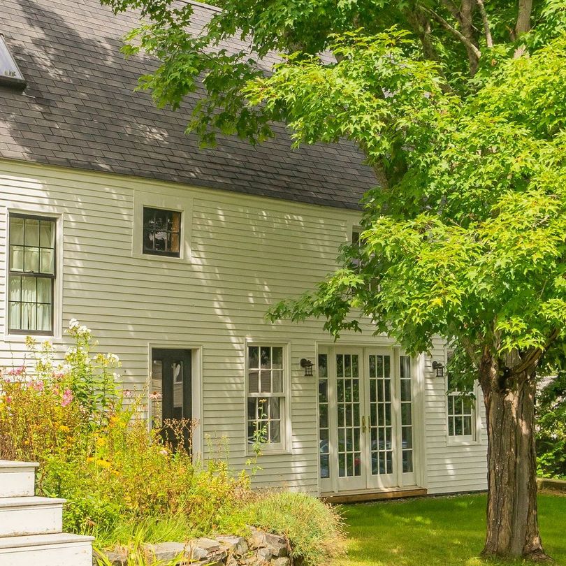 A white house with a black roof is surrounded by trees and bushes.