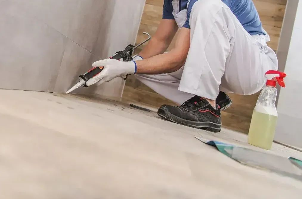 A person in white overalls caulking a tile joint with a caulk gun in a bathroom.