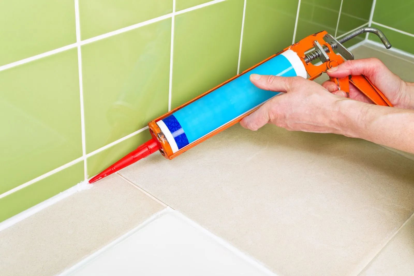 Person caulking grout in a bathroom. Green and tan tiles; orange caulk gun.
