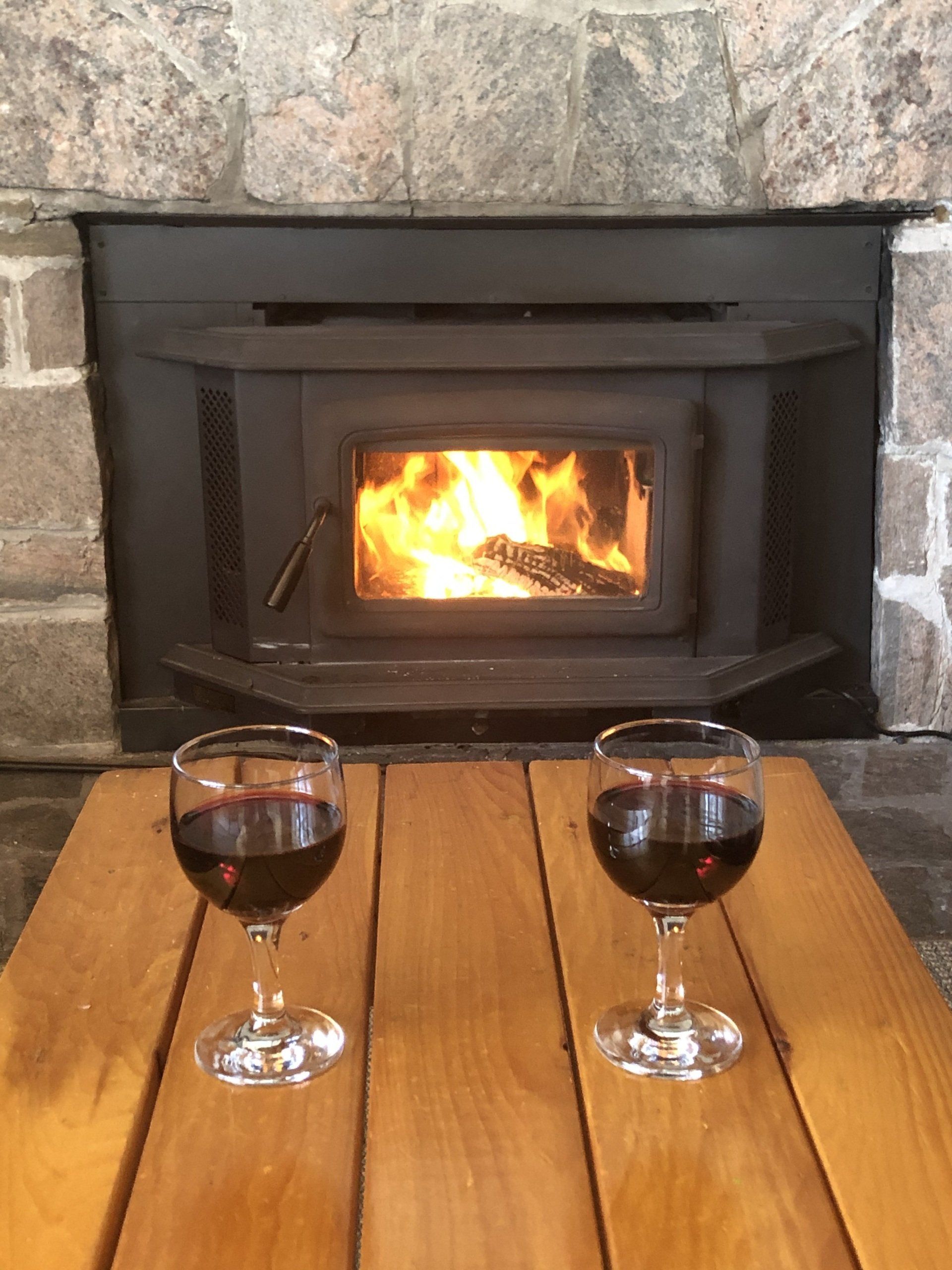 Two glasses of red wine are sitting on a wooden table in front of a fireplace.
