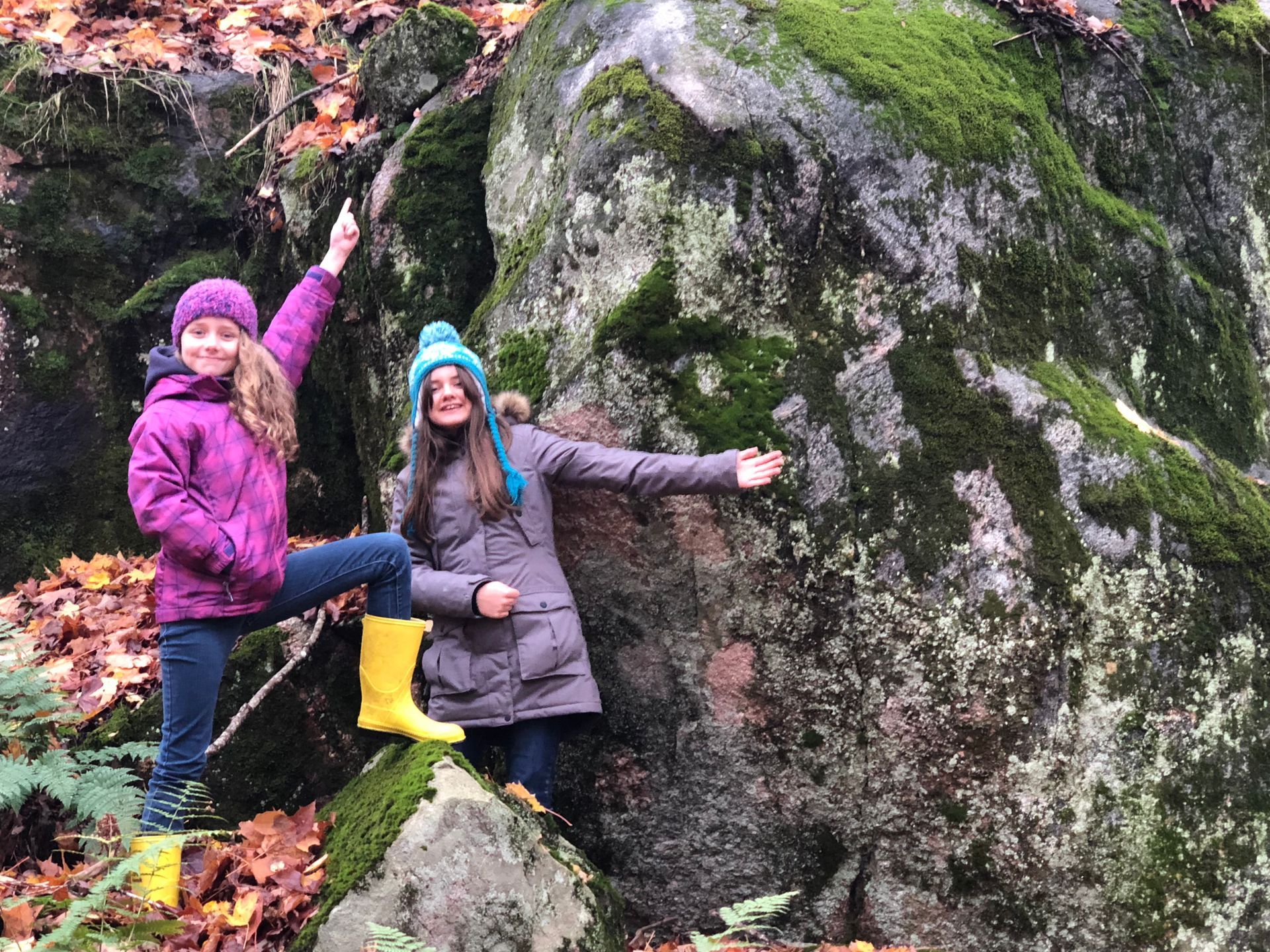 Two girls are standing next to a large rock in the woods.