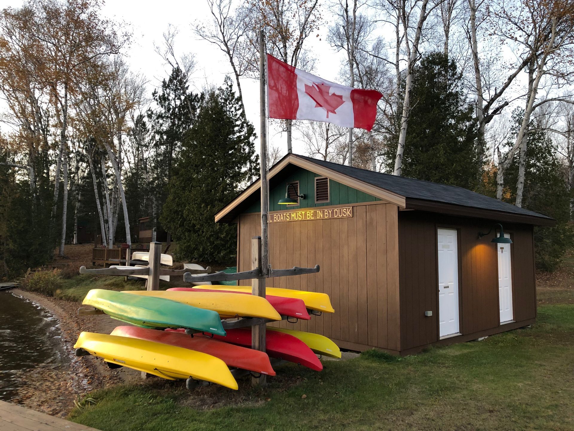 A row of kayaks are lined up in front of a shed with a canadian flag flying in the background.