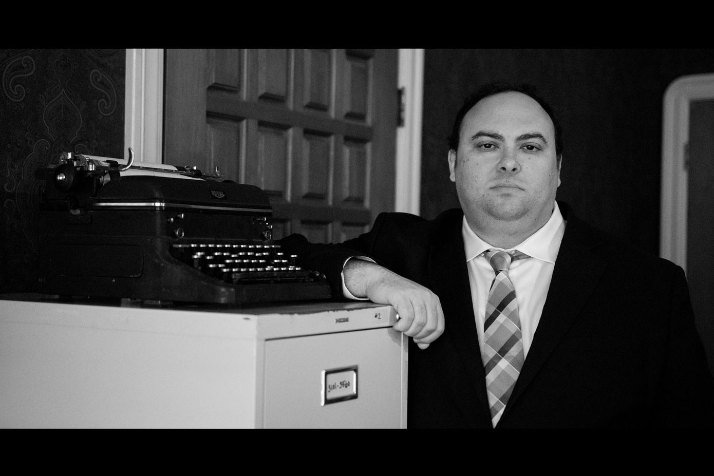 A man in a suit and tie is leaning on a filing cabinet next to a typewriter.