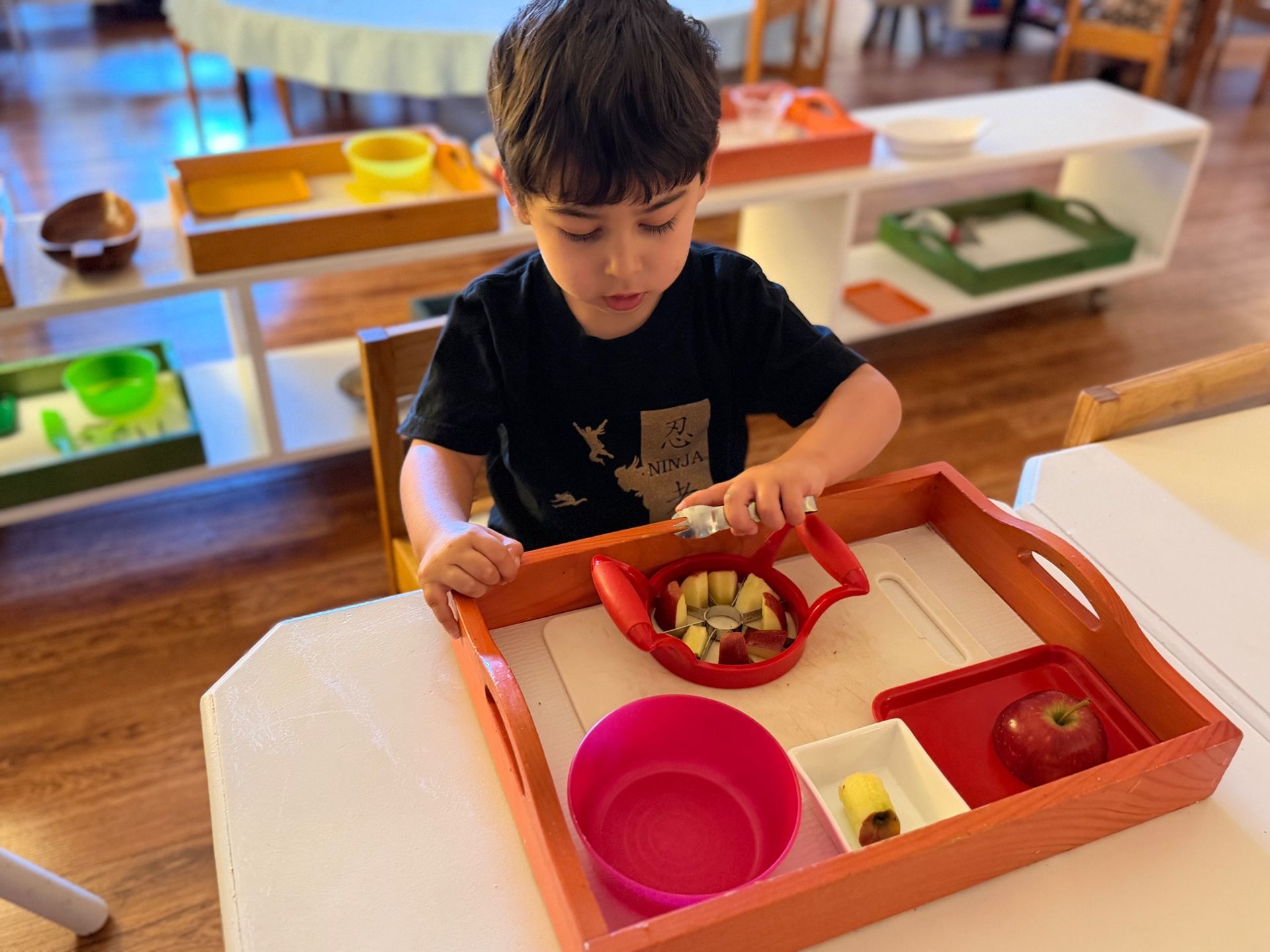 Child playing with toy food in a classroom, sorting items into colorful bins at a table.