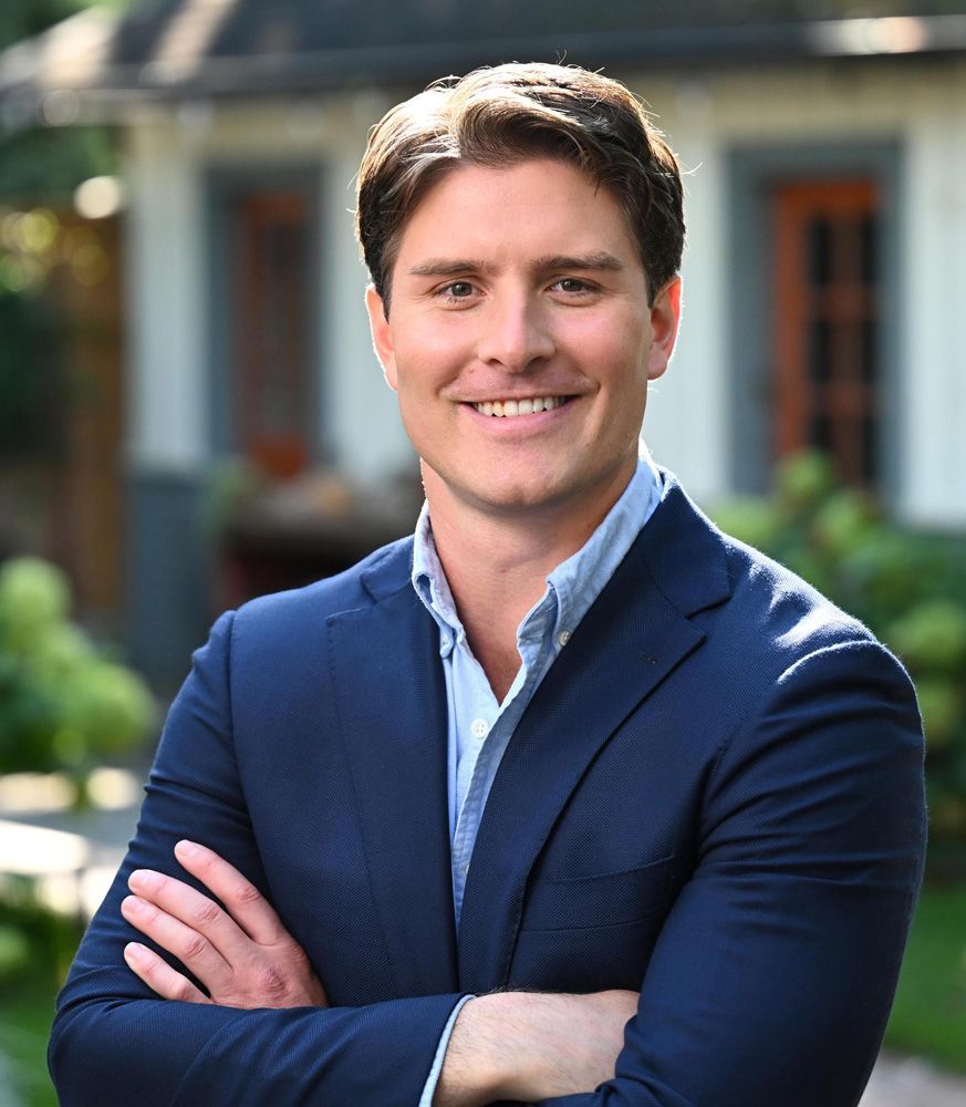 Man in a navy blazer and open light blue shirt, arms crossed, smiling.
