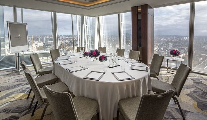 Uma sala de conferências com mesa oval, cadeiras e janelas do chão ao teto que oferecem uma vista panorâmica da cidade.