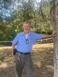 Man in blue shirt and green pants leans against a tree in a sunny outdoor setting.