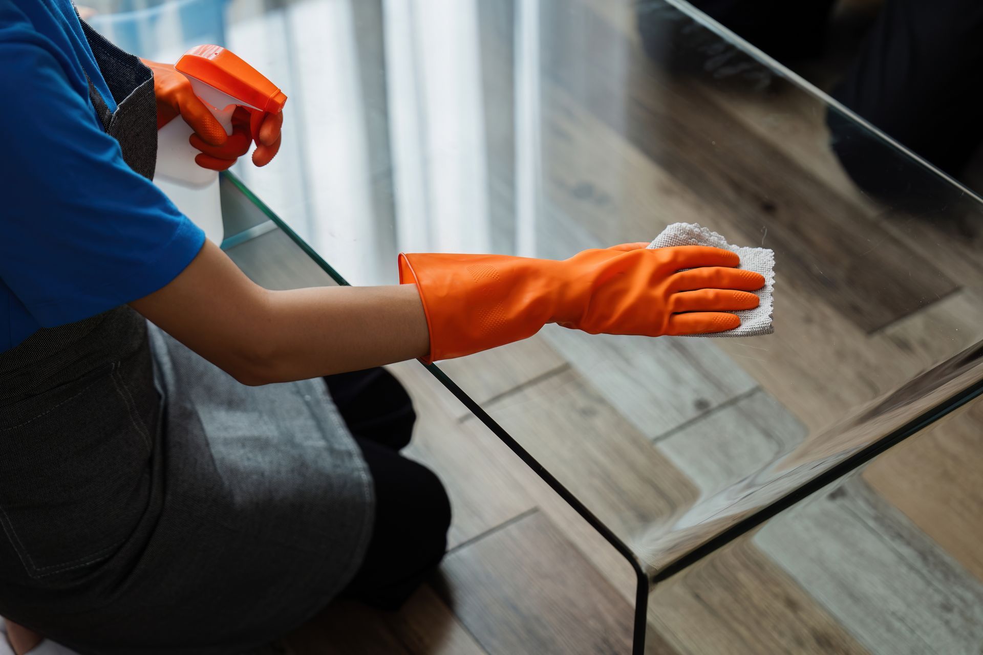 Person wearing orange gloves, spraying and wiping a glass table with a white cloth; indoor setting.