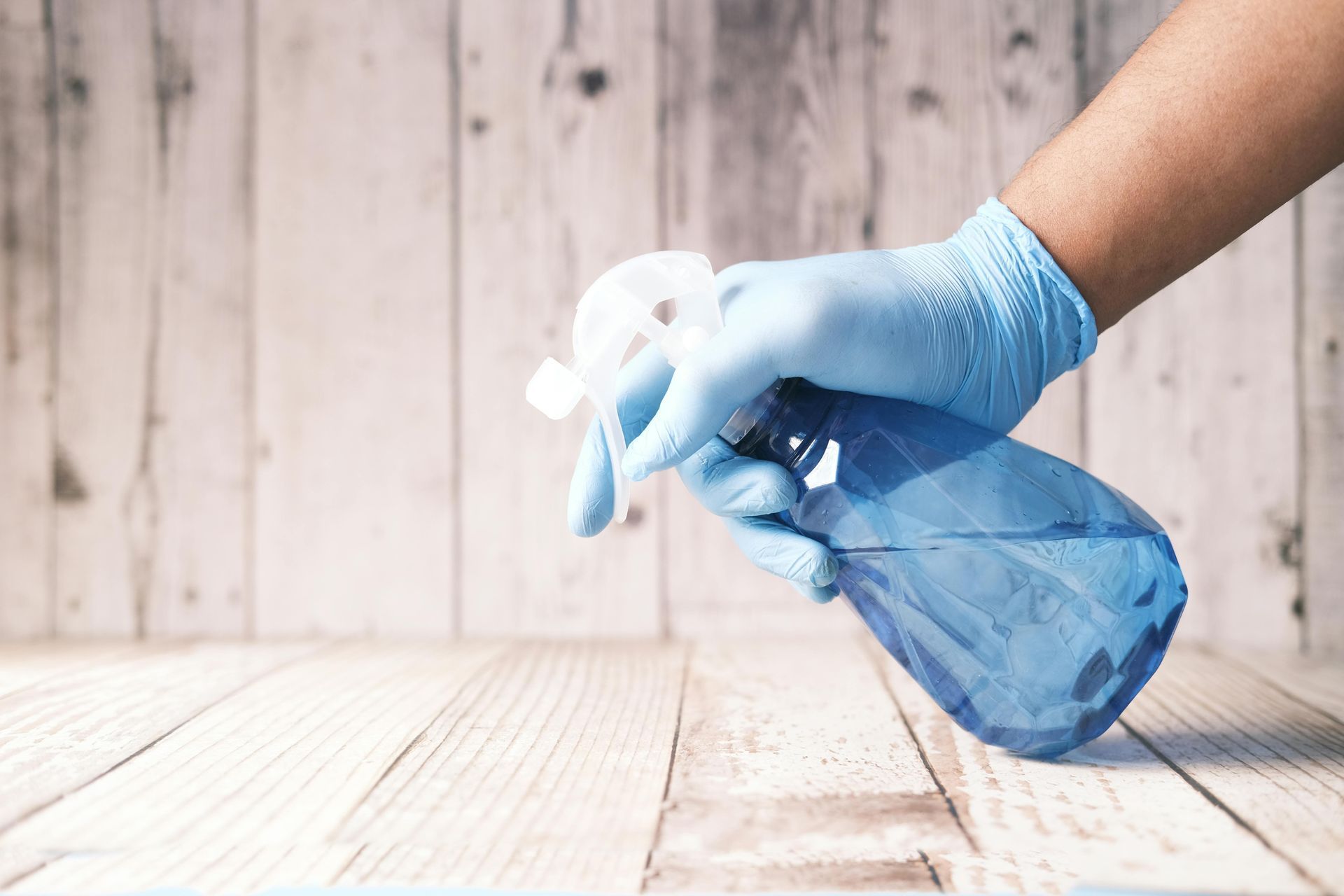 A person wearing blue gloves is cleaning a wooden floor with a spray bottle.