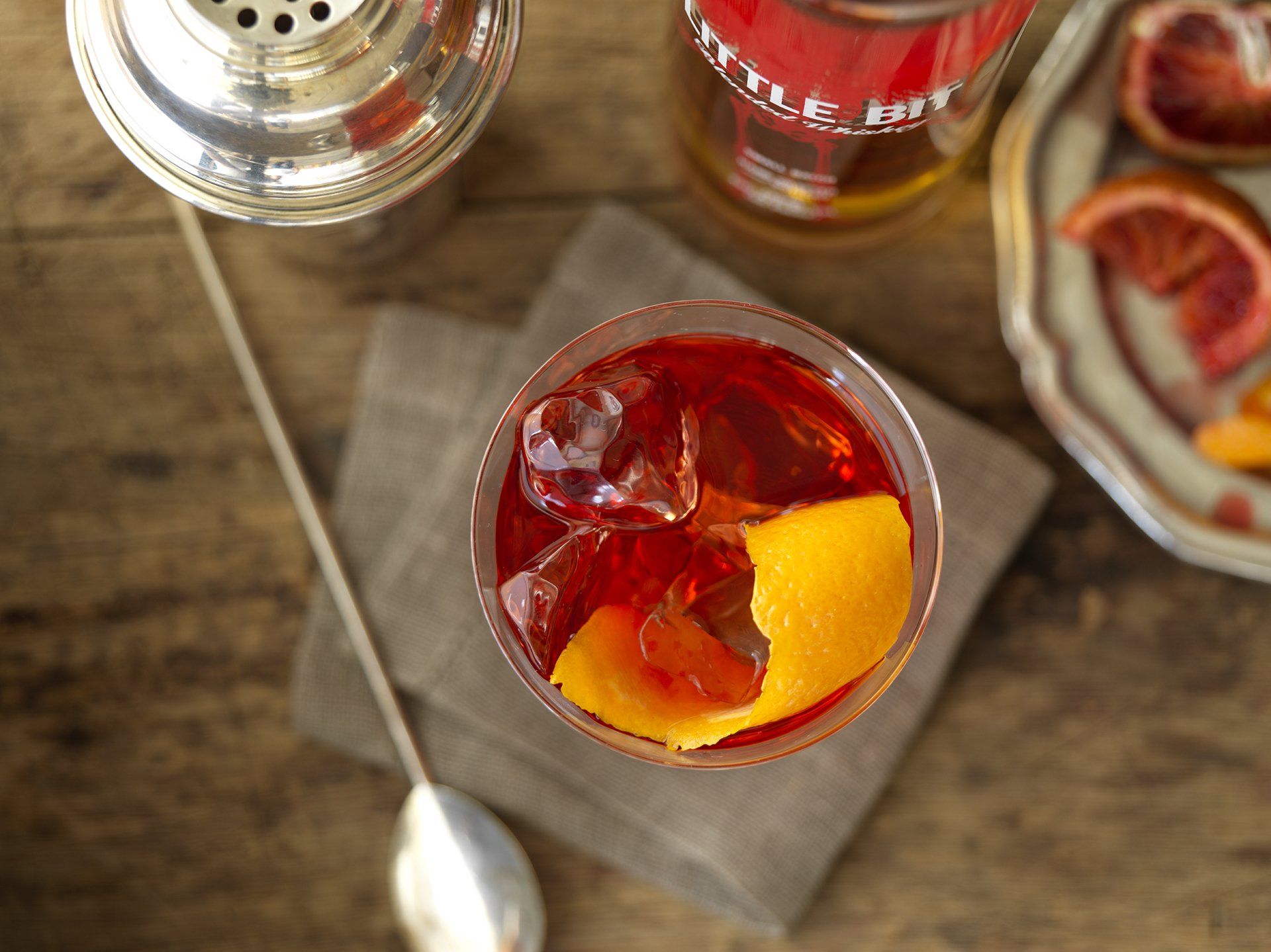 A glass of red liquid with ice and an orange peel on a wooden table.