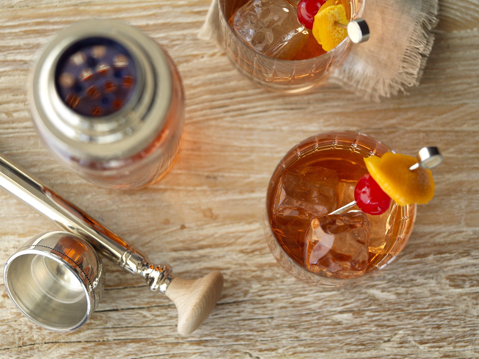A wooden table topped with two glasses of whiskey and a shaker.