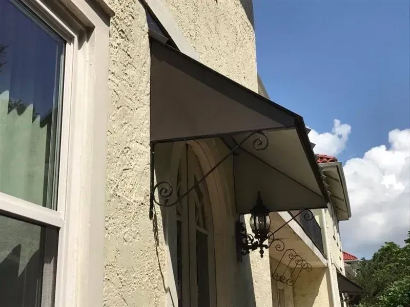 An awning over a window on a stucco building. It is dark gray with decorative metal supports.