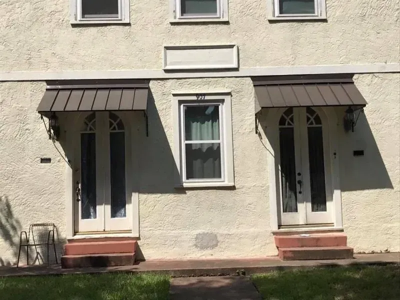 Two-story building exterior with two doors, awnings, and a central window. Beige stucco facade with brown accents and steps.