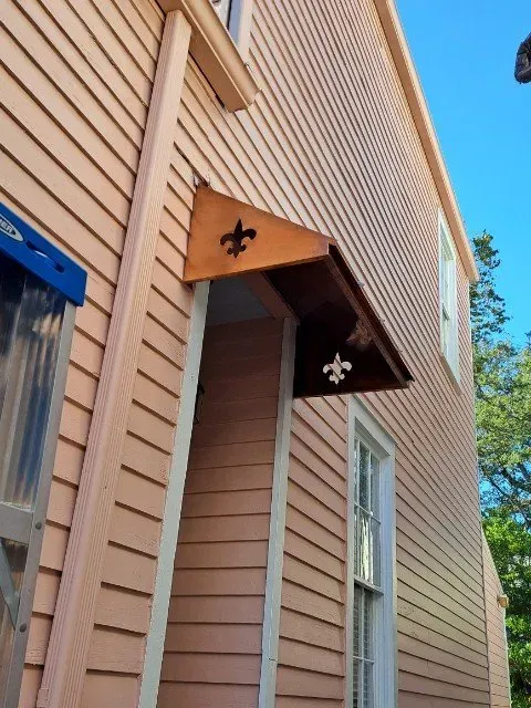 Peach-colored house with a dark wooden awning above a doorway, featuring a fleur-de-lis.