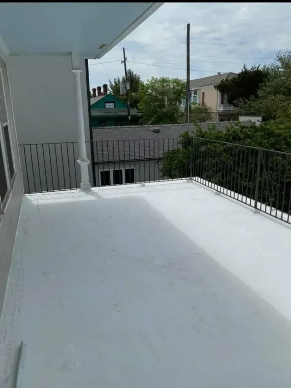 Balcony with white surface, black railing, view of houses and trees. Overcast sky.