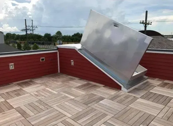 Rooftop access hatch, open. Silver metal hatch on red building with wood deck, cloudy sky background.
