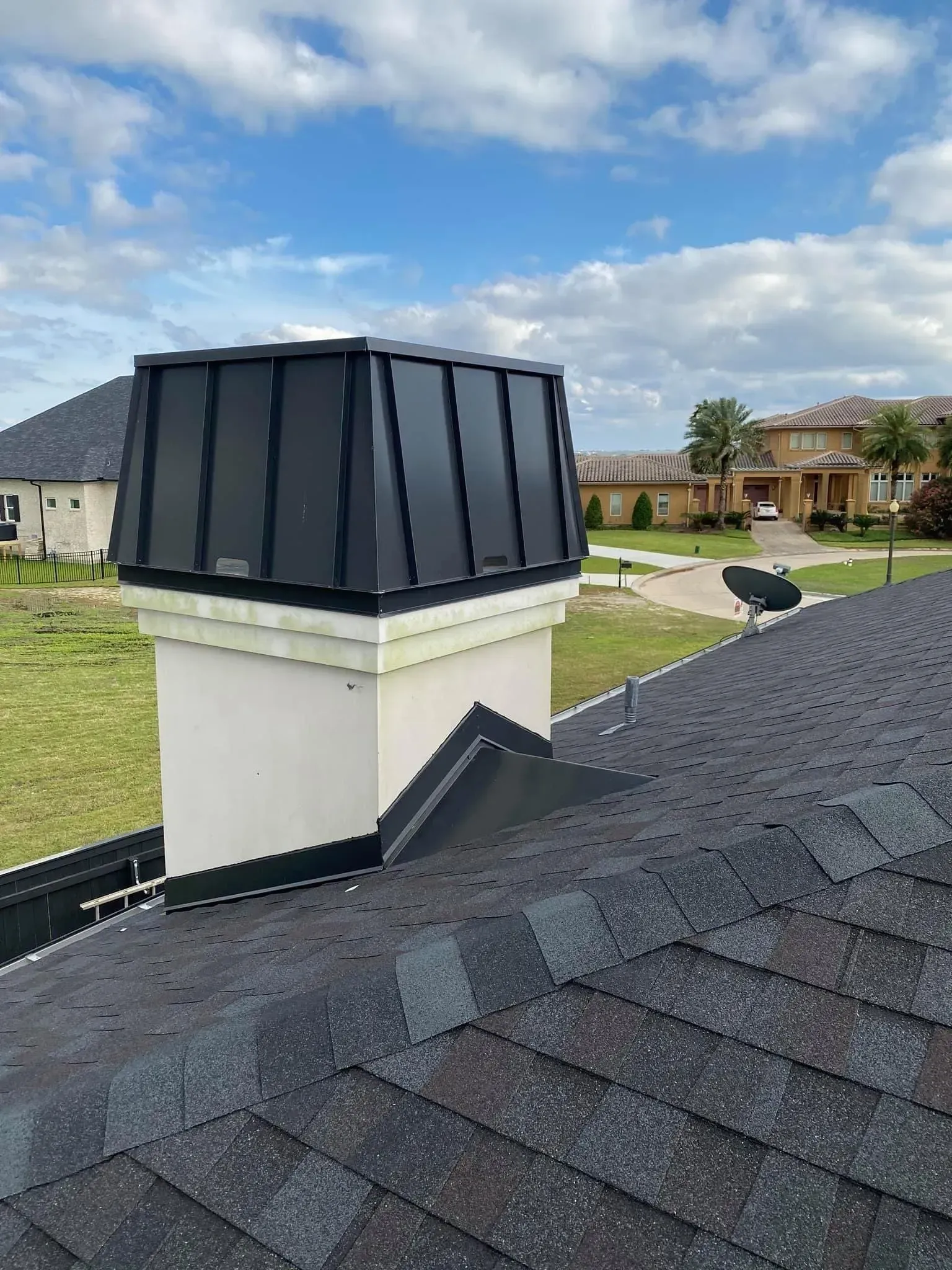 Chimney with black metal cap on a shingle roof against a cloudy sky.