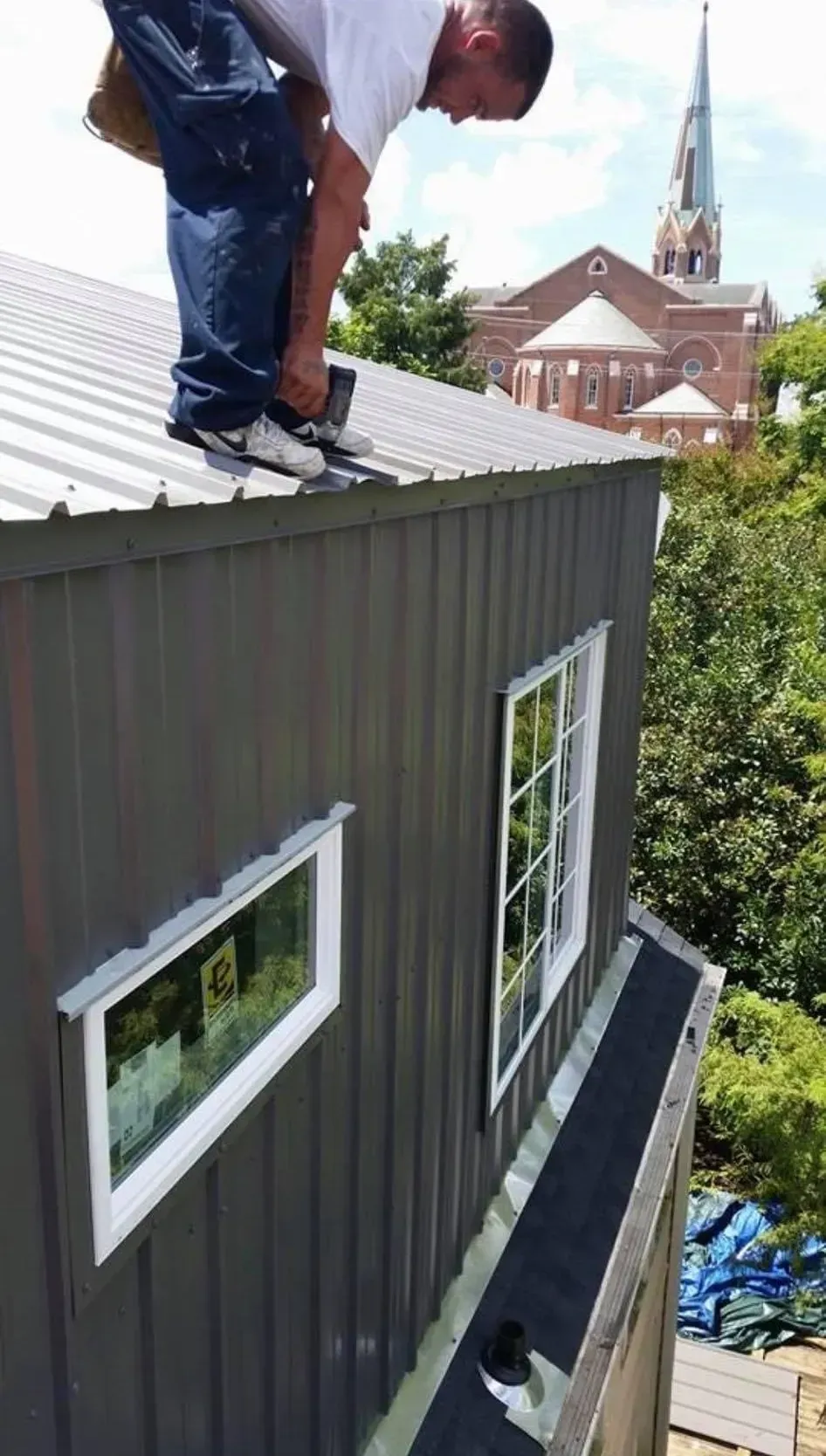 Person on a metal roof near two windows. Church and trees in the background. Gray building with white-framed windows.