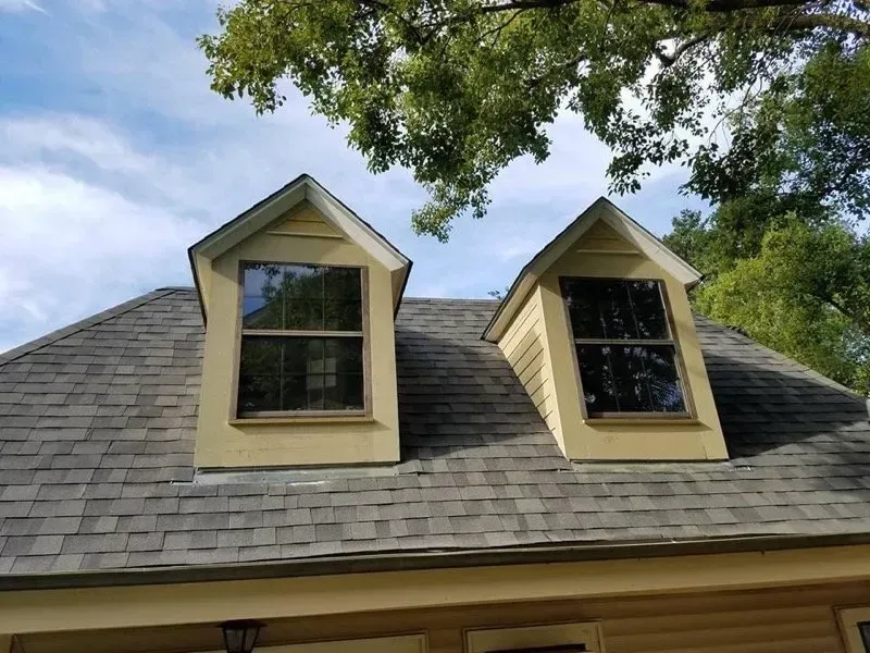 Two dormer windows on a gray-shingled roof. The windows are beige with dark glass, under a blue sky.