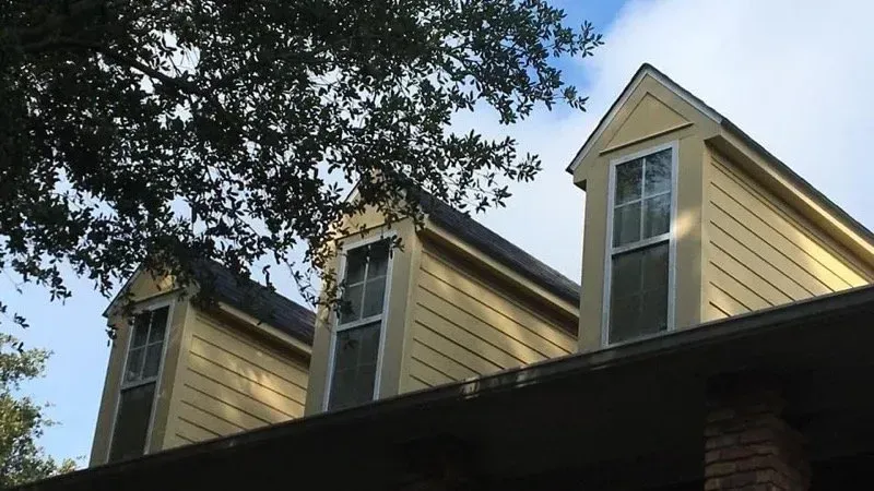 Three yellow dormer windows with white frames on a house roof, cloudy sky background.