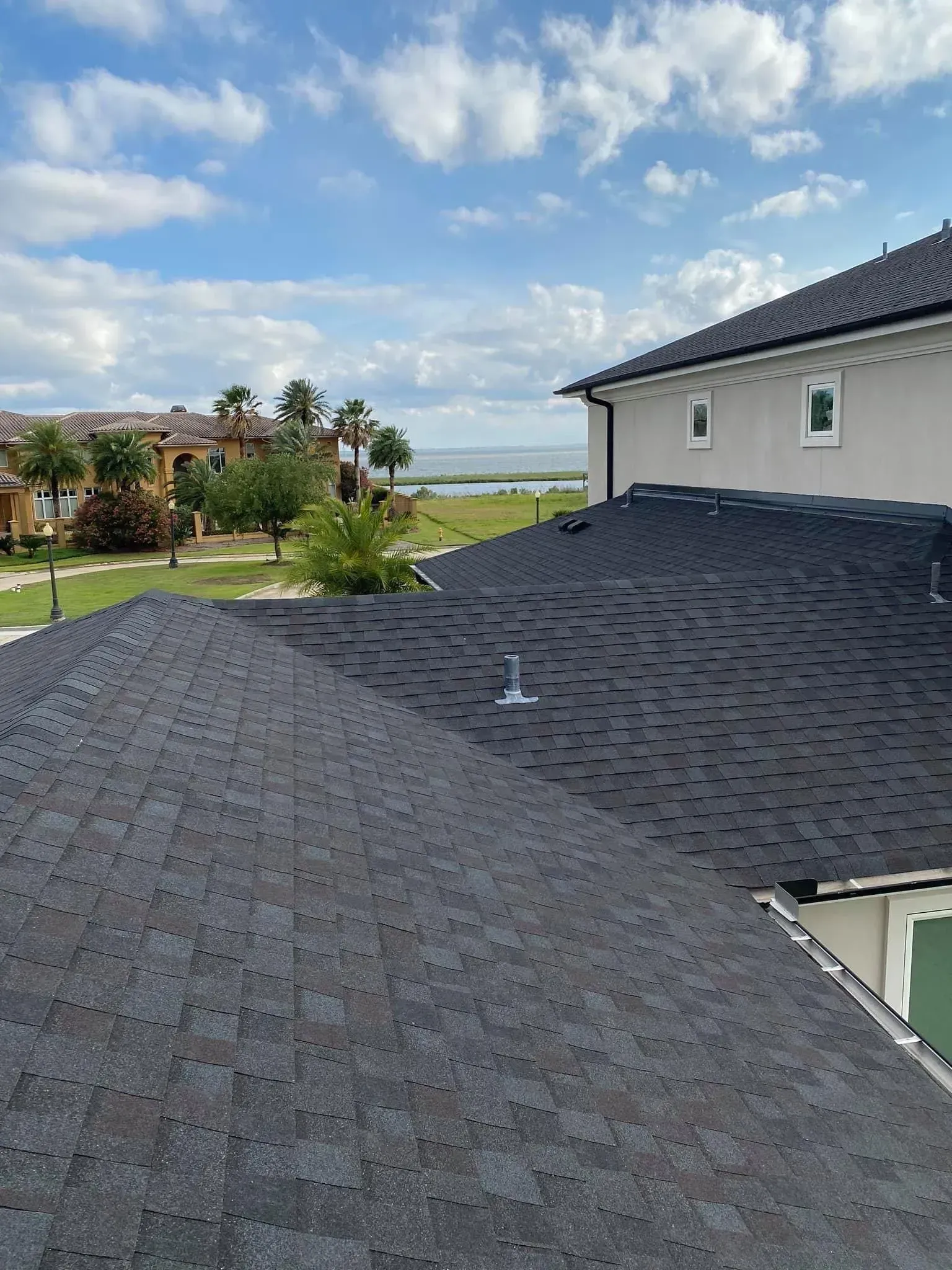 Dark asphalt shingle roofs on a house, with green grass, water, and blue sky in the background.