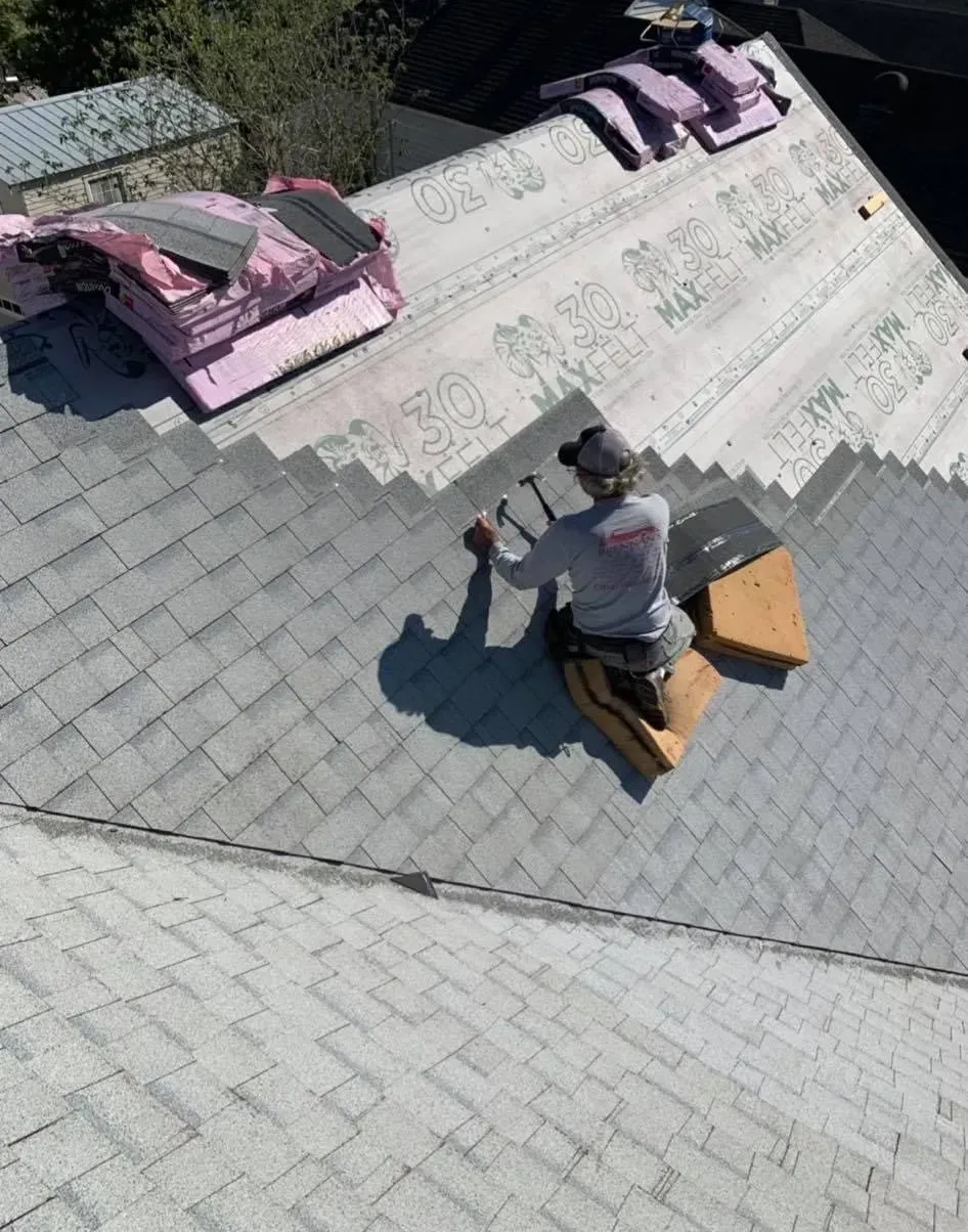 Roofer installing shingles on a house roof. Supplies and insulation are nearby. Sunny day.
