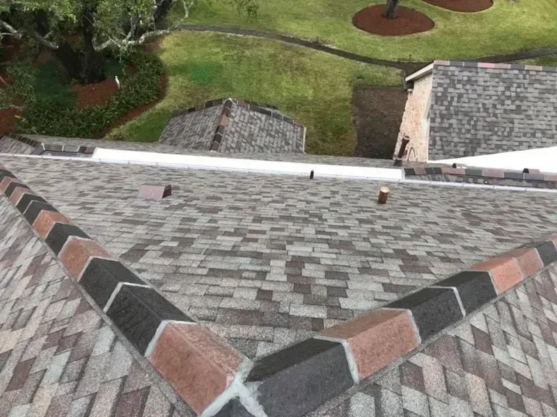 View of a brown and tan shingle roof with black and brick-colored trim, with a grassy yard in the background.