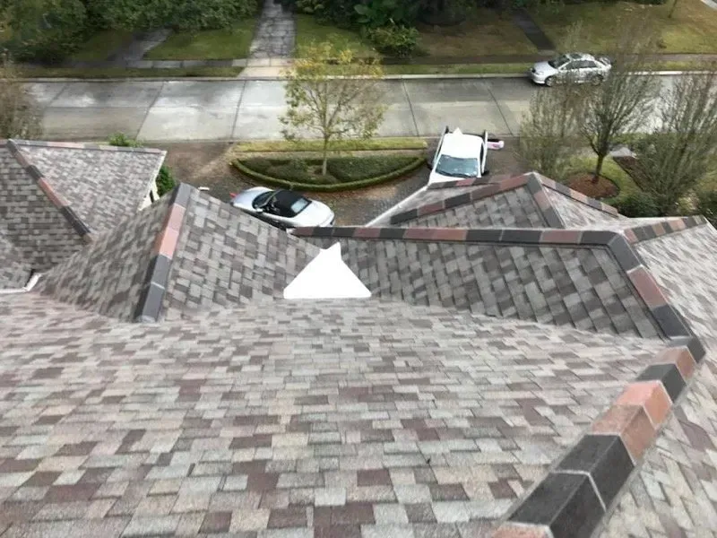 Overhead view of a house roof with multicolored shingles, a white vent, and a street view with cars.