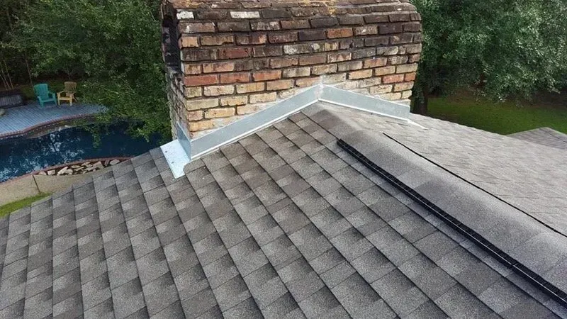 Brick chimney on a gray shingled roof with flashing, near a pool and trees.