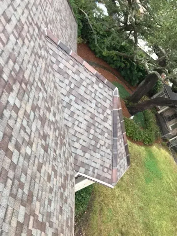 Close-up of a roof with brown and gray shingles, viewed from above, with trees and grass in the background.