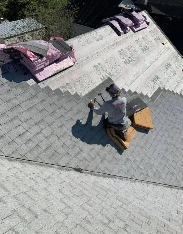 Roofer on a gray shingled roof, hammering shingles. Pink insulation and roof material are nearby. Sunny day.