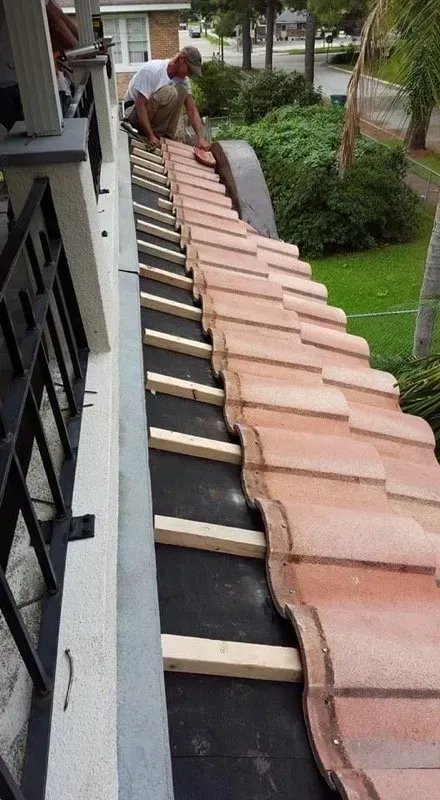 Man installing clay roof tiles on a house. Black underlayment, wooden supports, and a grassy yard are visible.