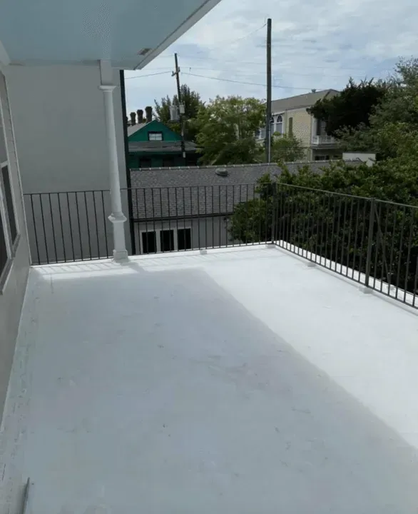 A white balcony with black railings overlooks a neighborhood with houses and trees under a cloudy sky.