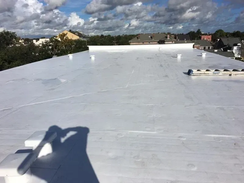 White flat commercial roof with vents and nearby buildings visible under a cloudy sky.