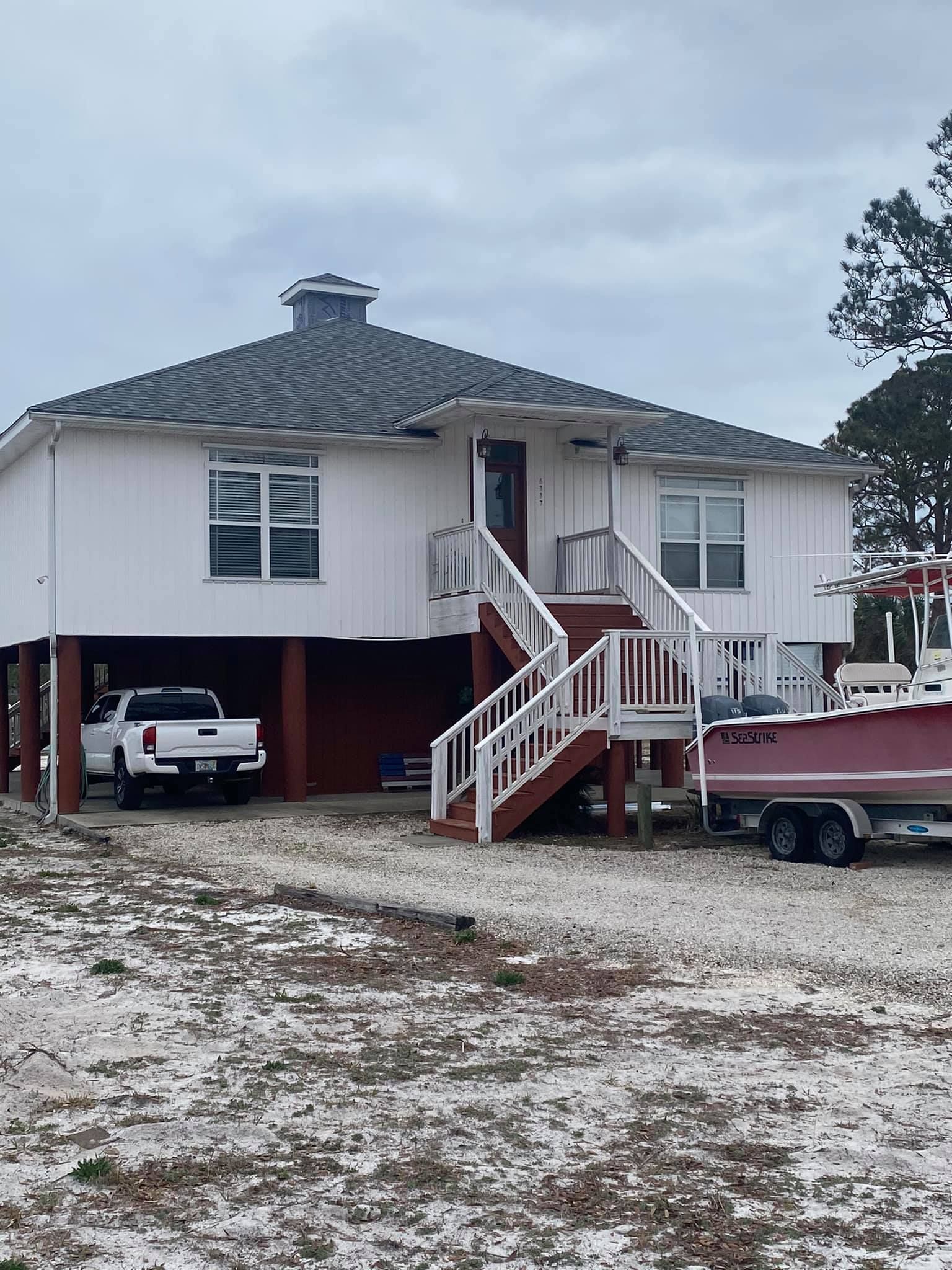 White house on stilts with a truck parked underneath. Boat on a trailer beside it. Cloudy day.