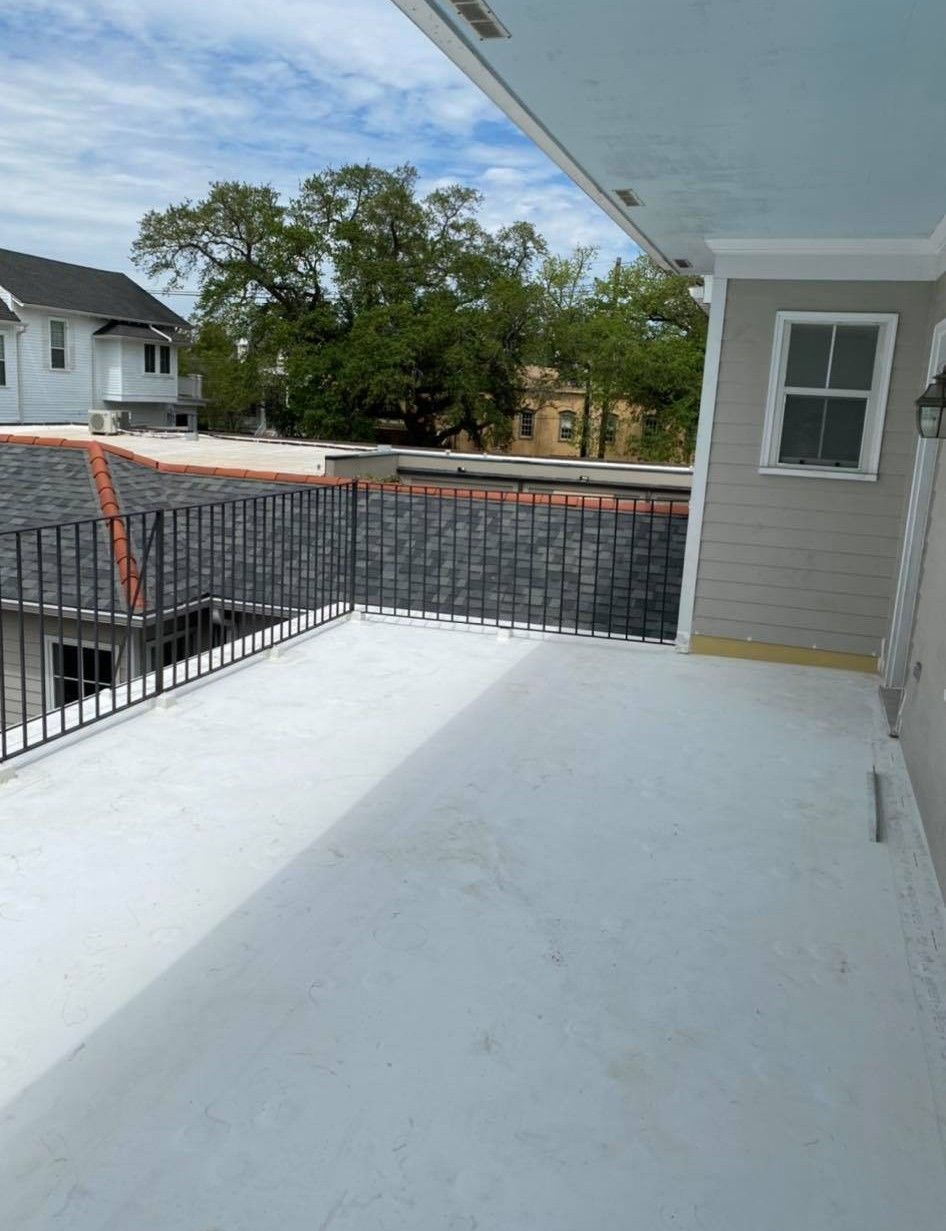 Rooftop deck with white surface, black railing, and grey siding. Buildings and trees in the background.