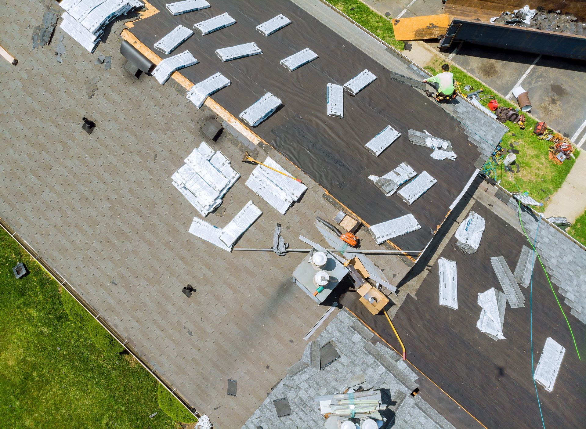 Aerial view of a roof under construction; workers, shingles, materials, and tar paper visible on the flat surface.