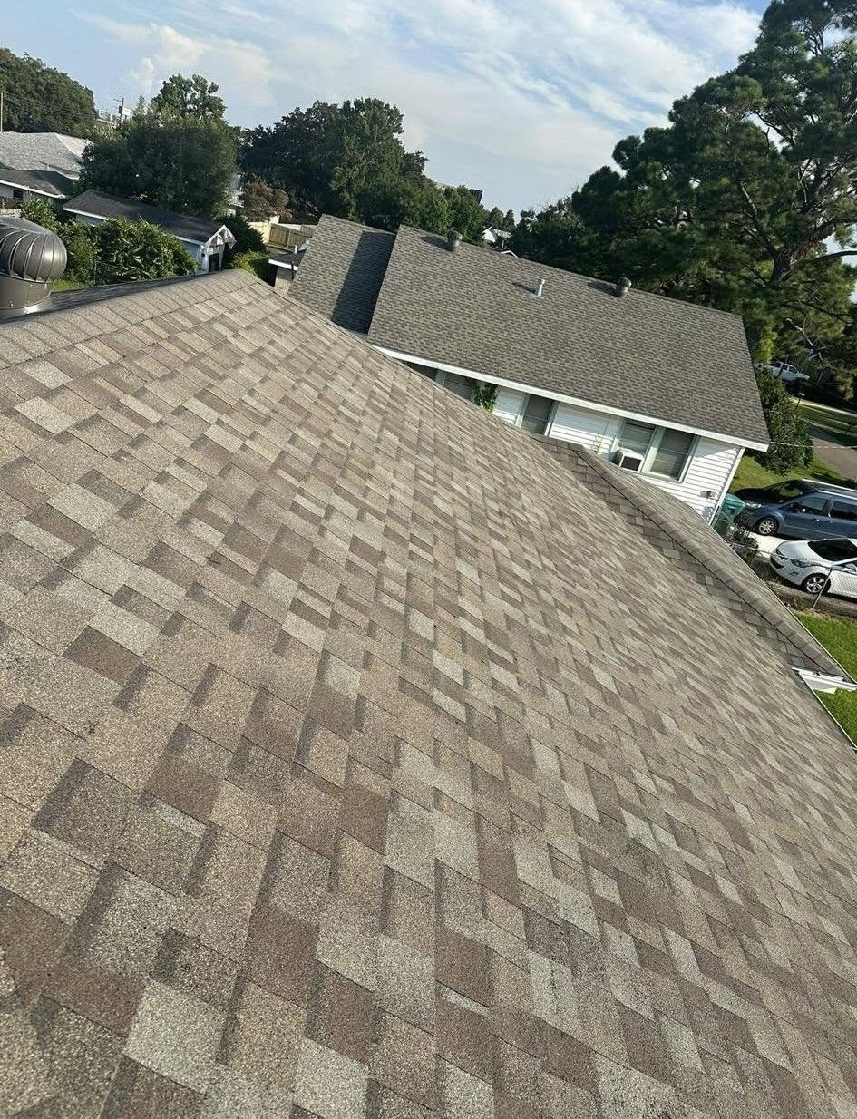 Overhead view of a roof with brown and gray asphalt shingles. Another roof and houses are in the background.