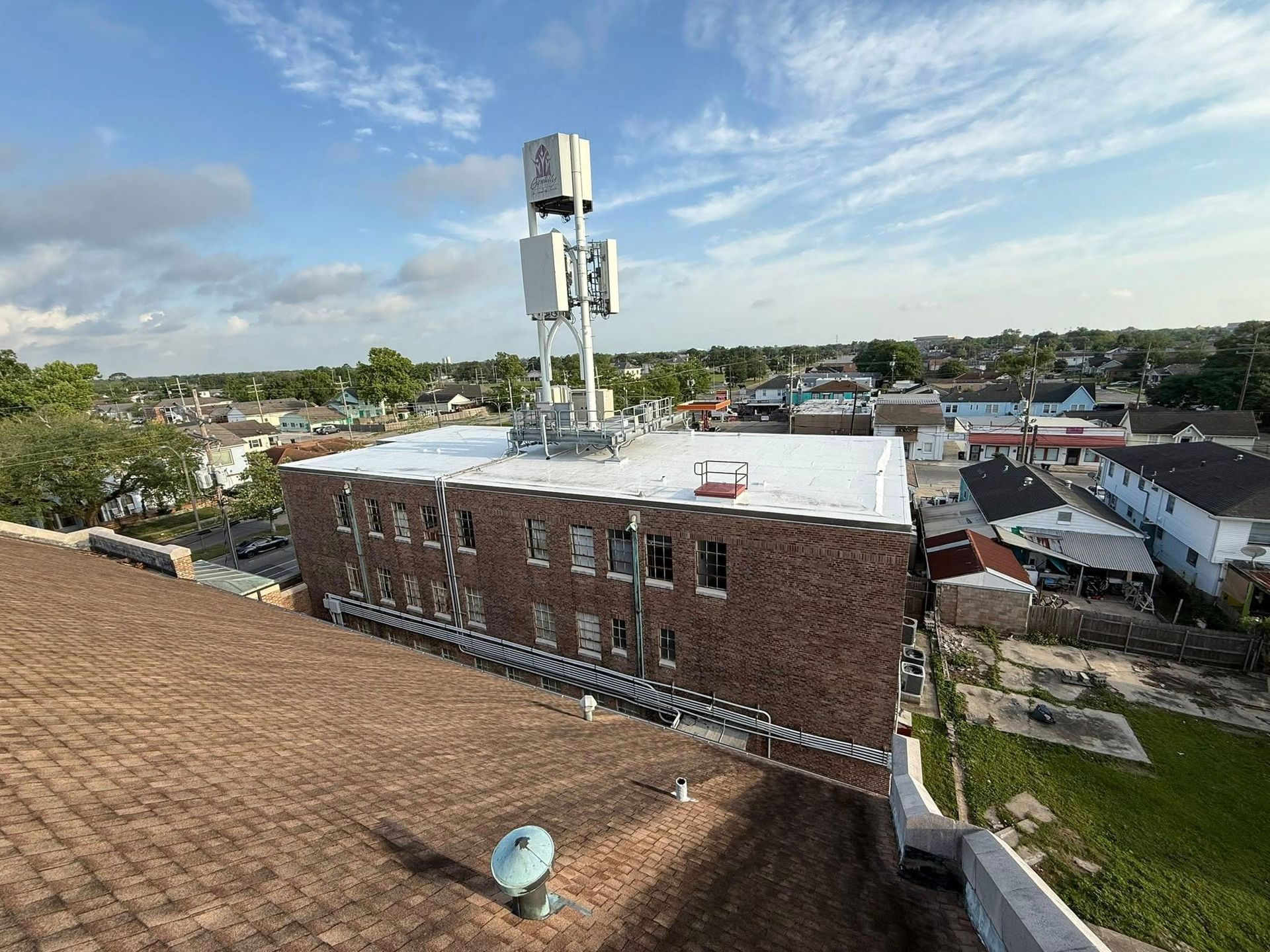 A brick building with a cell tower on the roof, surrounded by other buildings and a partly cloudy sky.