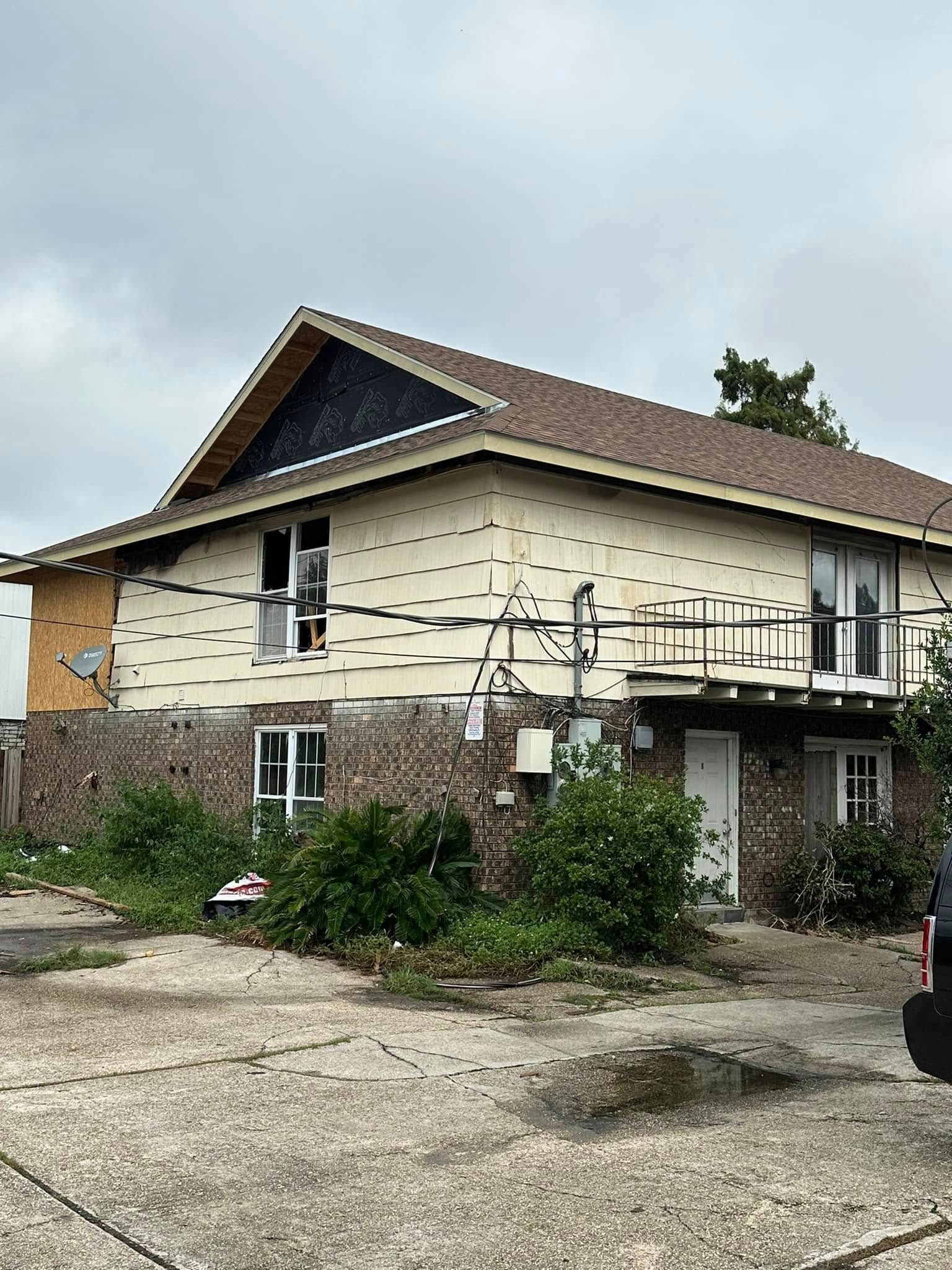 Two-story beige house with a brown roof and a small balcony. Exterior needs repairs, overgrown landscaping.