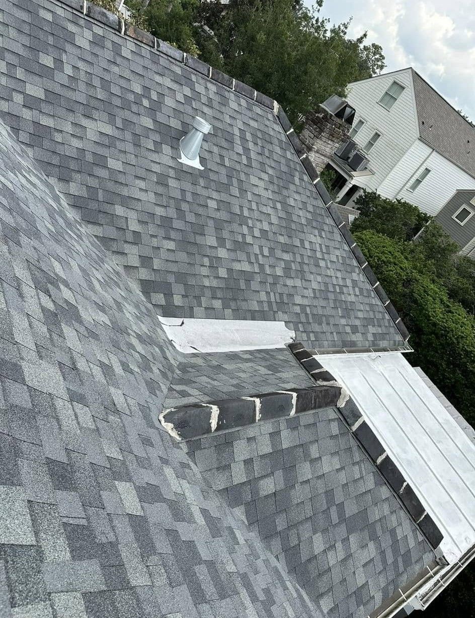 Gray asphalt shingle roof with a vent and a flat section. A house is in the background.