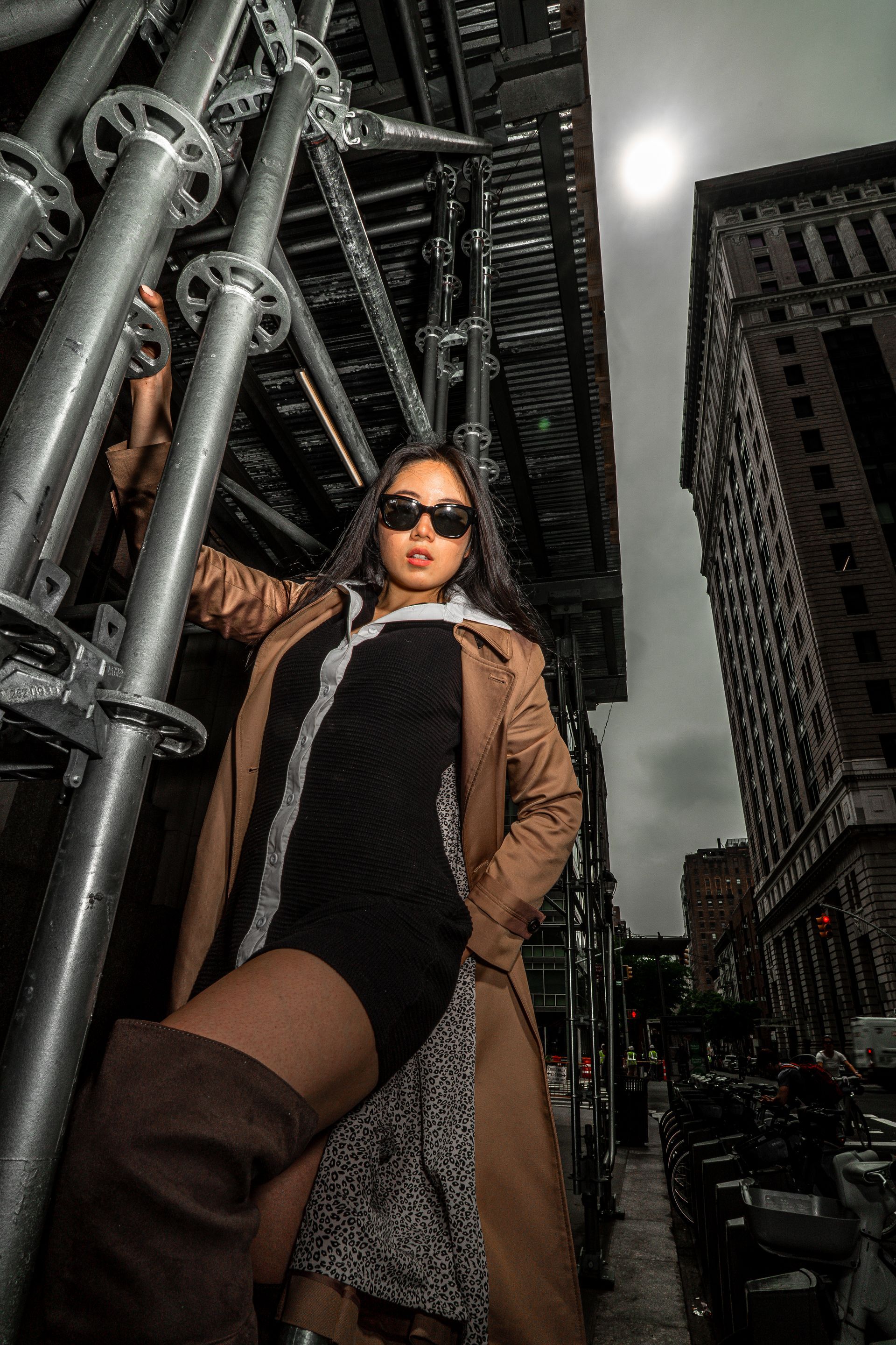 Woman in sunglasses poses by scaffolding; city background. Brown trench coat, black dress, tall boots.