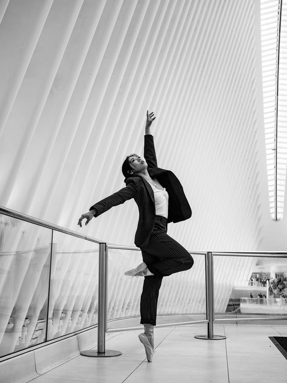Ballerina in a suit on pointe, poised in a modern, white building with large arches.