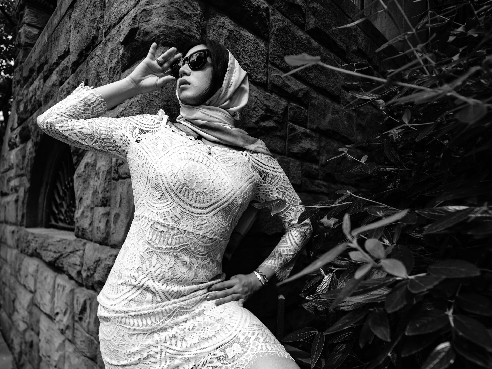 Woman in vintage looking white lace dress and sunglasses and white scarf poses outside near stone wall, hand to forehead.