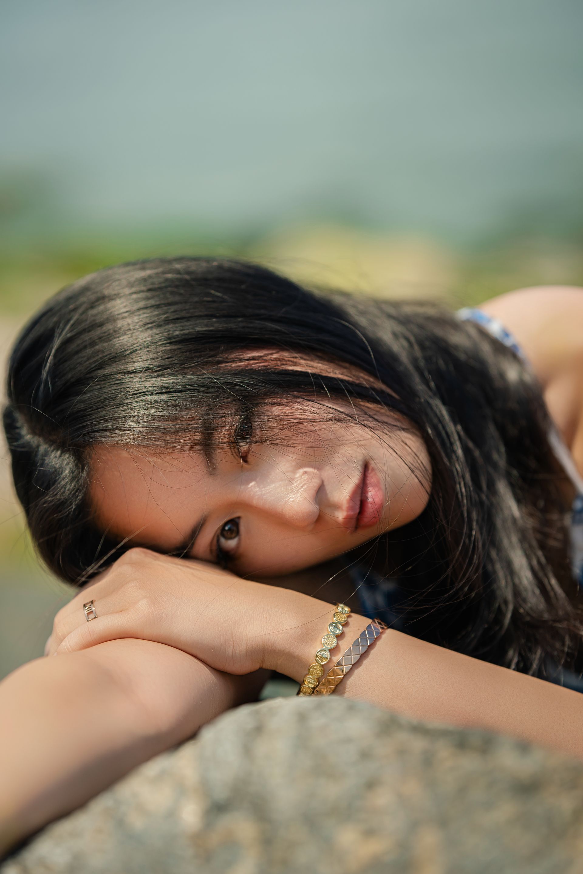 NYC model Minnie Lee — resting head on a rock, looking at the camera. Long dark hair, wearing a blue dress, outdoors. Fashion editorial photographed by Alexis Pesante